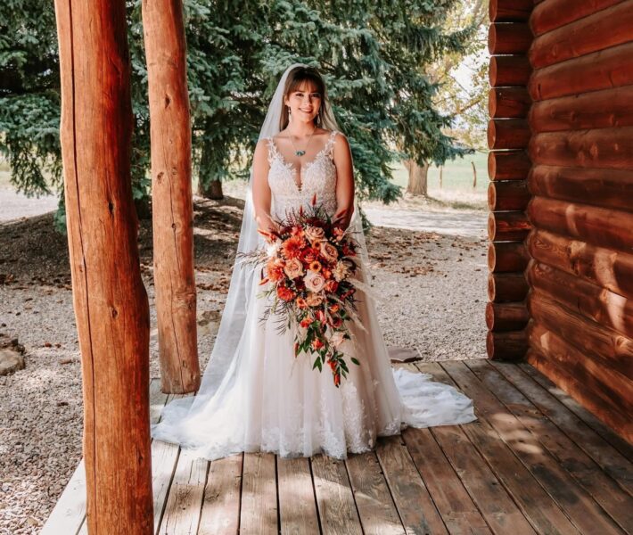 Bride in a stunning lace wedding dress holding a vibrant bouquet of roses and greenery, standing on a wooden porch surrounded by nature and rustic log cabin elements.