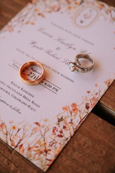 Wedding invitation featuring elegant floral designs with a gold wedding band and a diamond engagement ring placed on top. The invitation details the wedding date, September 27th, 2023, at Wrich Ranches in Crawford, Colorado.