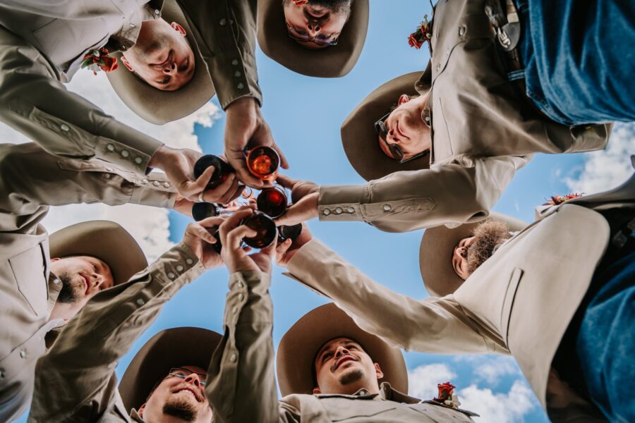 Group of men in beige suits and wide-brimmed hats celebrating with drinks, viewed from below against a blue sky with clouds. The scene captures camaraderie and festivity, ideal for themes of celebration, weddings, or gatherings.