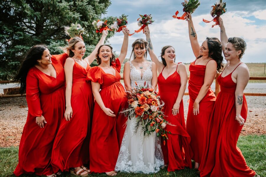 Bridal party celebrating outdoors, featuring the bride in a white lace gown and bridesmaids in vibrant red dresses, joyfully holding floral bouquets. The scene is set against a backdrop of greenery and a cloudy sky, capturing a moment of happiness and camaraderie.