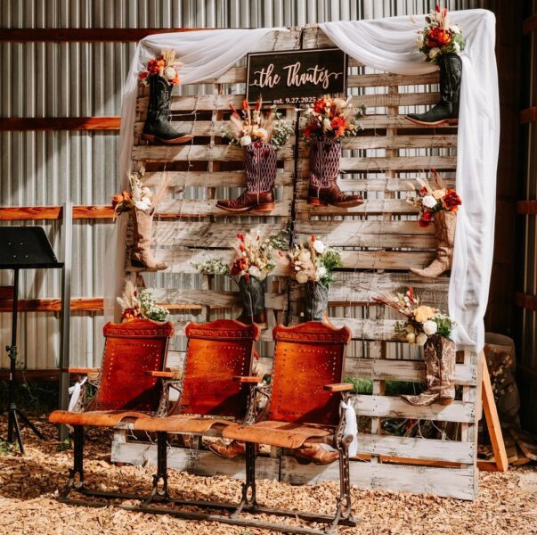 Rustic wedding decor featuring a wooden backdrop adorned with cowboy boots and floral arrangements, with three vintage wooden chairs in the foreground. The sign reads 