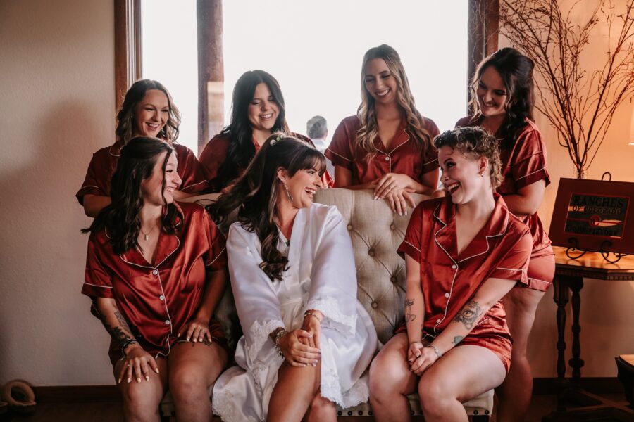 Bridal party in matching red satin pajamas joyfully celebrating together, with the bride in a white robe, seated in a cozy indoor setting. The image captures a moment of laughter and camaraderie among friends, perfect for wedding preparation themes.