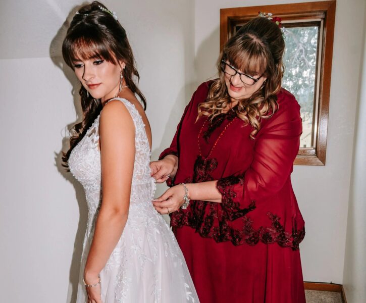 Bride getting ready for her wedding as her mother helps fasten the back of her intricately designed wedding dress, showcasing a moment of love and support in a well-lit room.