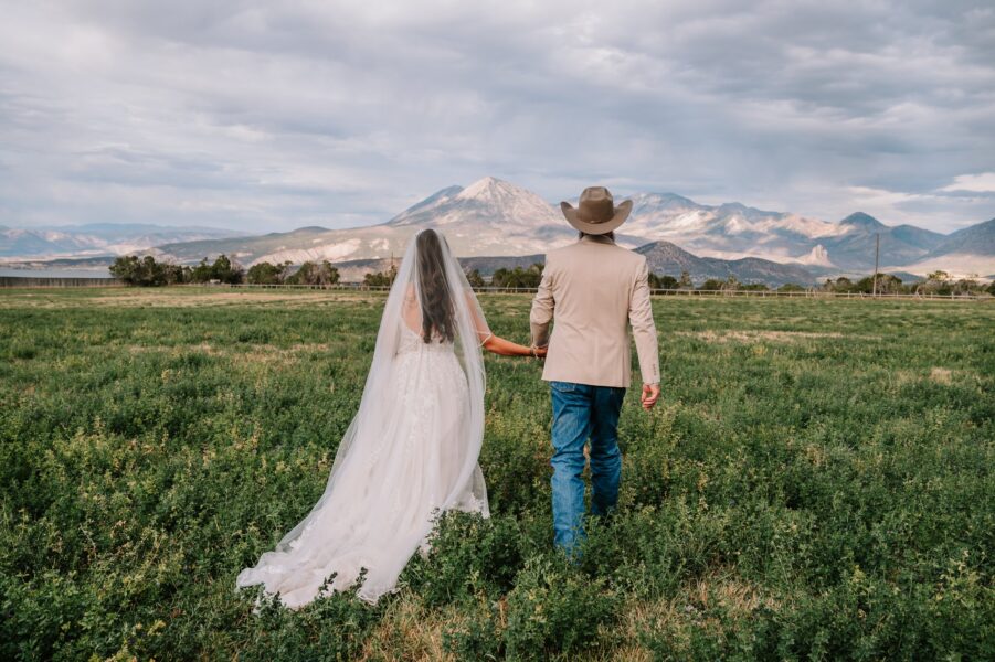 Bride and groom walking hand in hand through a lush green field, with majestic mountains in the background and a cloudy sky, capturing a romantic outdoor wedding moment.