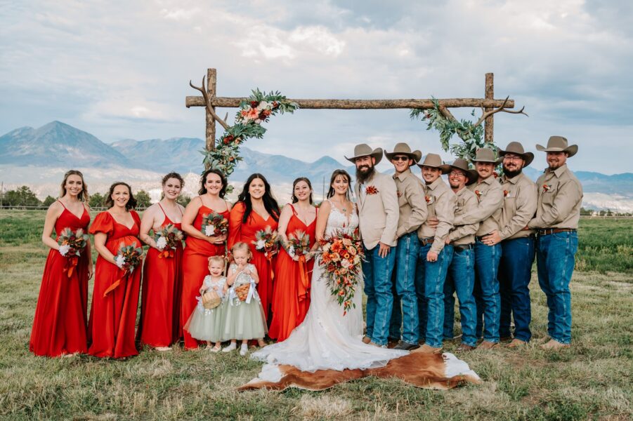 Bridal party in red dresses and cowboy attire posing together under a floral archway in a scenic outdoor setting with mountains in the background. The bride, wearing a white gown, stands in the center surrounded by bridesmaids and groomsmen, capturing a joyful wedding moment.