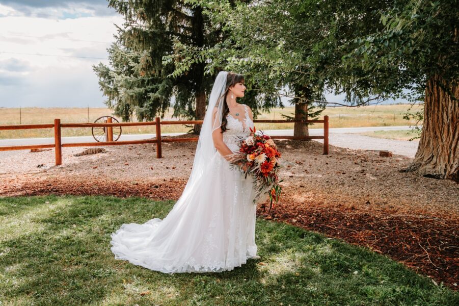 Bride in a white wedding dress holding a vibrant bouquet of flowers, standing outdoors in a picturesque setting with trees and a rustic wooden fence in the background.