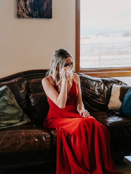 Young woman in a red dress sitting on a brown leather couch, holding a tissue to her face, with a scenic view visible through a window in the background.