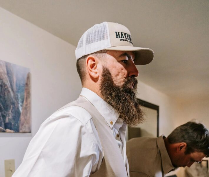 Profile of a man with a long beard wearing a baseball cap and a light-colored vest, standing in a room with soft lighting and a framed landscape picture on the wall. Another man is partially visible in the background, focusing on an activity.