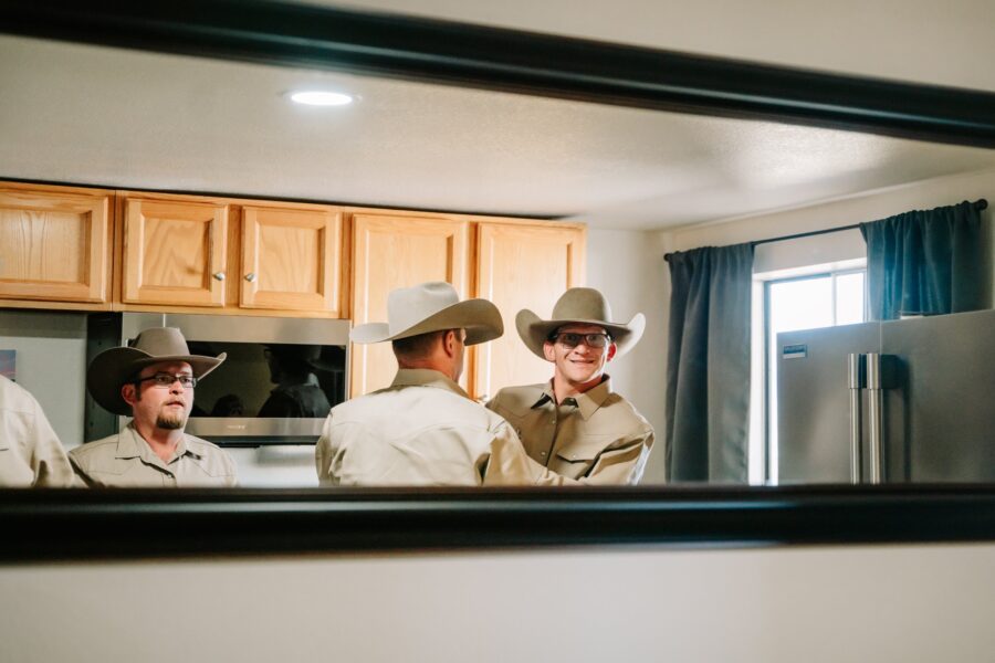 Three men in cowboy hats and matching shirts are interacting in a kitchen, reflected in a mirror. The kitchen features wooden cabinets and a stainless steel refrigerator, with natural light coming through a window.
