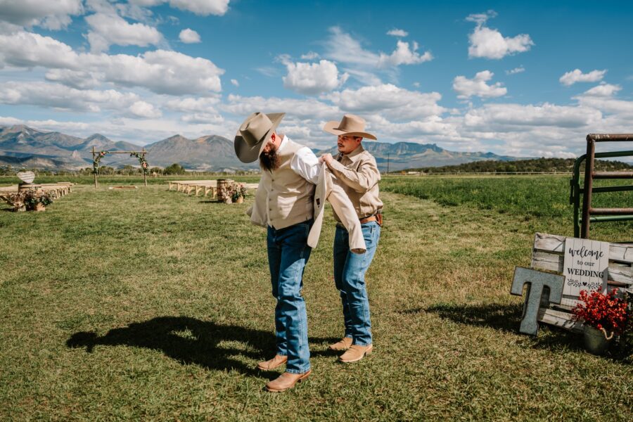 Two men prepare for a wedding in a scenic outdoor setting, with mountains in the background. One man helps the other put on a jacket while both are dressed in western attire, including hats and boots. A wooden sign nearby welcomes guests to the wedding.