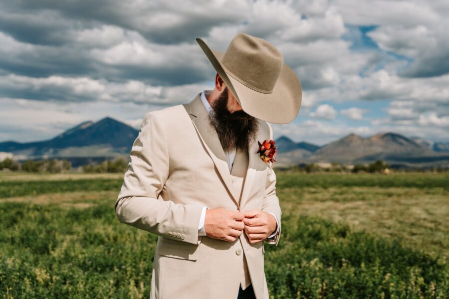 Groom adjusting his beige suit jacket with a large cowboy hat and floral boutonnière, set against a scenic backdrop of mountains and a cloudy sky.