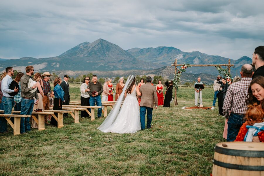 Outdoor wedding ceremony with a bride in a white gown walking down the aisle with her father, surrounded by guests on wooden benches. The backdrop features a scenic mountain landscape and an arch decorated with flowers. The wedding party includes bridesmaids in red dresses and a groom at the altar.