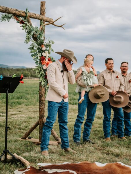 Groom emotional during outdoor wedding ceremony, standing in front of a floral-decorated wooden arch. Surrounded by groomsmen in casual attire and a young girl in a green dress, with a picturesque landscape in the background.
