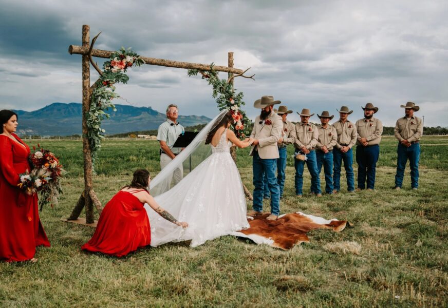 Outdoor wedding ceremony with a bride and groom exchanging vows under a floral arch, surrounded by groomsmen in cowboy attire and bridesmaids in red dresses. A scenic mountain backdrop enhances the rustic atmosphere.