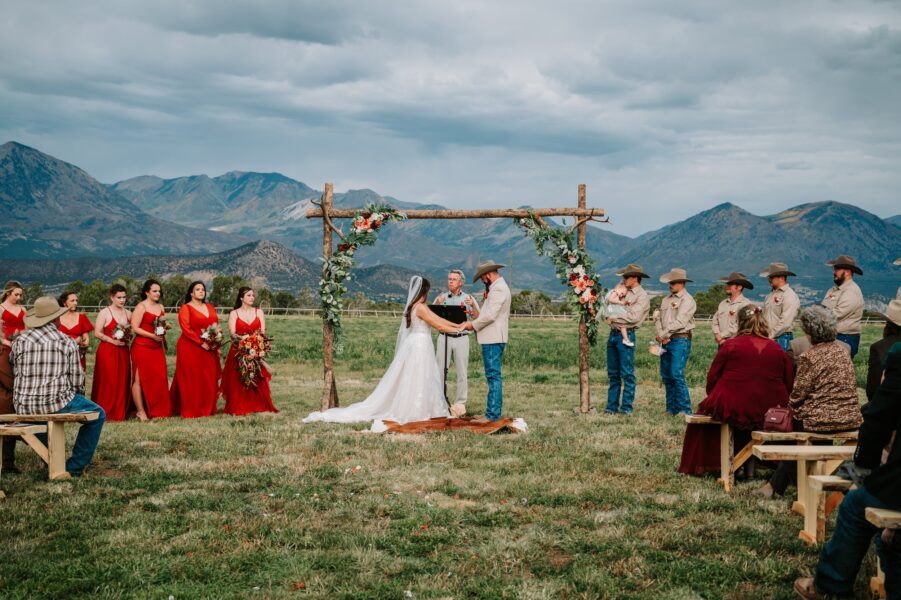 Outdoor wedding ceremony in a scenic mountain setting, featuring a bride and groom exchanging vows under a floral arch, surrounded by bridesmaids in red dresses and groomsmen in cowboy attire. Guests are seated on wooden benches, creating a rustic and intimate atmosphere.