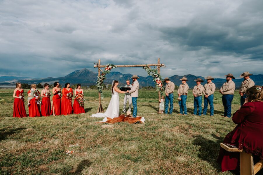 Outdoor wedding ceremony in a scenic mountain setting, featuring a bride in a white gown and a groom in a beige suit exchanging vows under a floral arch. Bridesmaids in red dresses and groomsmen in cowboy attire stand nearby, with a young girl and guests observing the event.