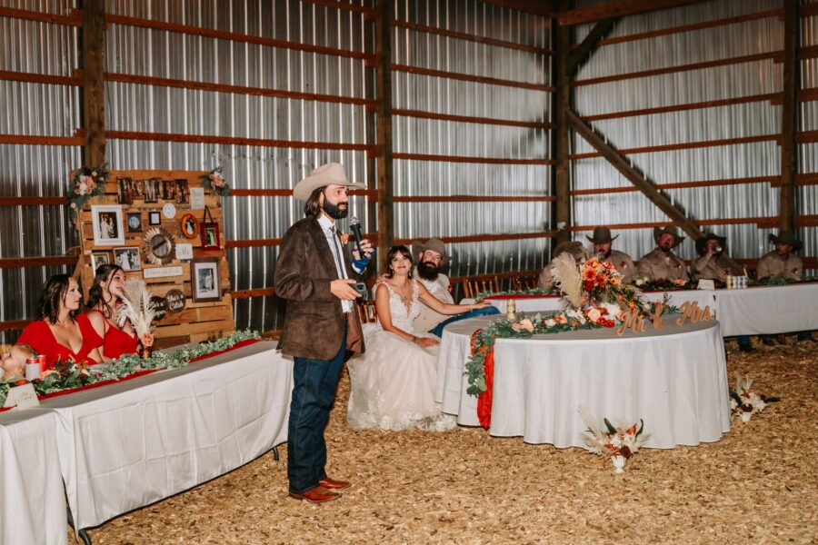Groom giving a speech at a rustic wedding reception in a barn, with the bride seated beside him. Guests in formal attire are seated at tables decorated with flowers and greenery, while a photo display is visible in the background.
