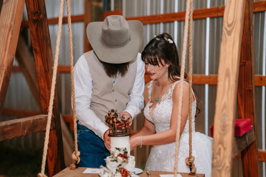 Bride and groom in rustic setting, engaged in a cake-cutting ceremony. The bride, wearing a lace wedding dress and delicate jewelry, and the groom, dressed in a vest and wide-brimmed hat, are focused on a beautifully decorated wedding cake adorned with floral accents. The background features wooden beams and a cozy atmosphere, emphasizing a romantic and intimate celebration.