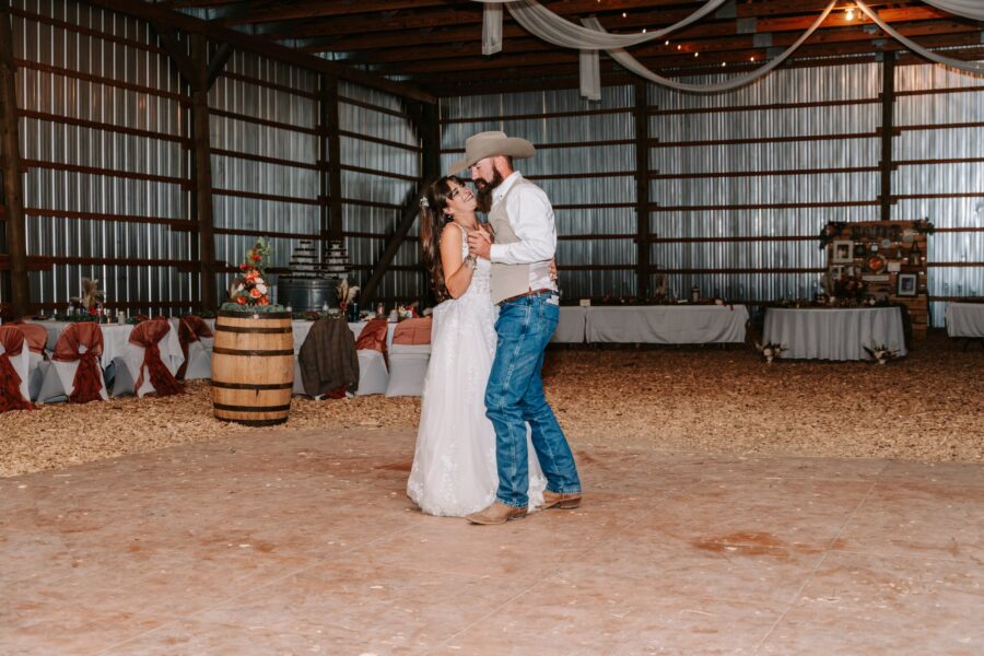 Couple dancing romantically at a rustic wedding reception in a barn, surrounded by decorated tables and wooden accents. The bride wears a white lace gown while the groom is dressed in a cowboy hat and casual attire, creating a charming country atmosphere.