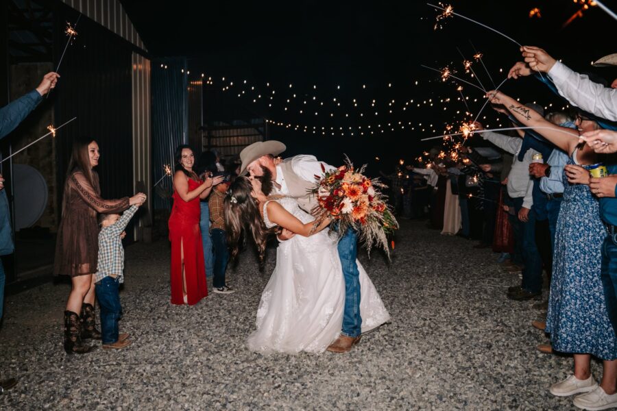 A joyful wedding celebration scene featuring a couple sharing a romantic kiss as guests hold sparklers, creating a festive atmosphere. The bride, in a white dress, holds a vibrant floral bouquet, while the groom, wearing a cowboy hat and vest, leans in for the kiss. Guests of all ages, including children, are dressed in country-style attire, with twinkling lights in the background enhancing the celebratory mood.