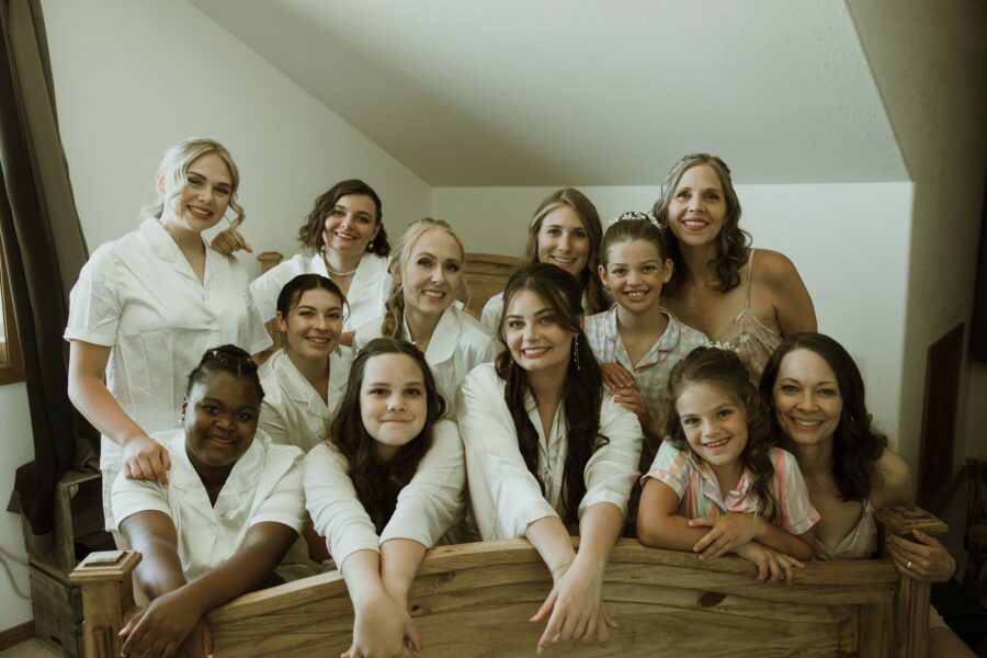 A joyful group of bridesmaids and young girls in matching white and pastel outfits, posing together in a cozy indoor setting, celebrating a wedding day with smiles and laughter.