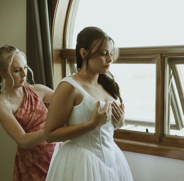 Bride adjusting her wedding dress with the help of a bridesmaid by a window, showcasing a moment of preparation before the ceremony.