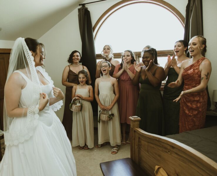 Bridal party celebrating with the bride in her wedding dress, showcasing joyful reactions in a beautifully lit room. The group includes women and girls in various elegant dresses, capturing a heartfelt moment before the ceremony.