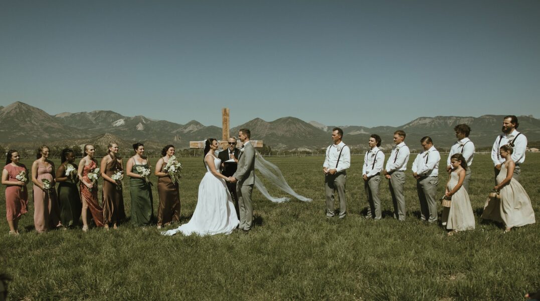 Bride and groom exchanging vows during an outdoor wedding ceremony in a scenic mountain setting, surrounded by bridesmaids and groomsmen in coordinating attire, with a wooden cross in the background.