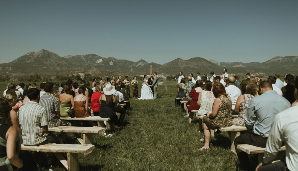 Outdoor wedding ceremony with guests seated on wooden benches, set against a backdrop of mountains and clear blue sky. The couple exchanges vows at the altar, surrounded by floral arrangements, creating a picturesque and romantic atmosphere.