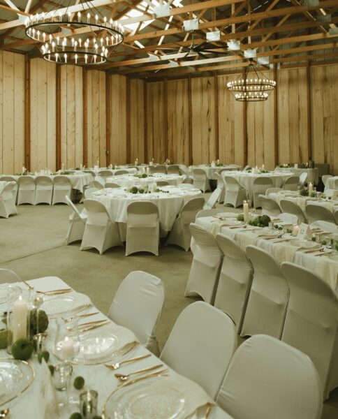 Elegant banquet hall setup featuring round tables with white linens and gold accents, surrounded by white chairs. The space is illuminated by large chandeliers and showcases a rustic wooden interior, perfect for weddings or events.