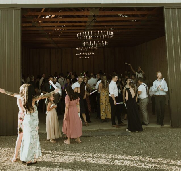 Guests dancing and celebrating inside a rustic venue with chandeliers, enjoying a lively atmosphere during a wedding reception, with some holding glow sticks and wearing formal attire.