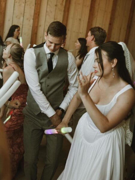 A joyful wedding celebration scene featuring a couple dancing with glow sticks, surrounded by guests in a rustic venue with wooden walls. The bride is in a white dress, while the groom wears a gray suit. The atmosphere is lively and festive, capturing the essence of a fun wedding reception.
