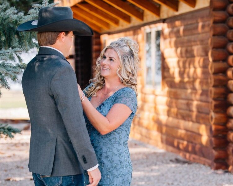 Couple sharing a joyful moment outdoors, with a rustic log cabin in the background. The woman is wearing a blue dress with intricate patterns, while the man is dressed in a gray blazer and cowboy hat, creating a warm, romantic atmosphere.