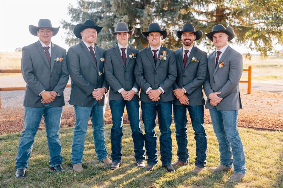 Group of six men in gray suits and cowboy hats, standing together outdoors with a rustic background. Each man is wearing a boutonniere, showcasing a stylish western-themed attire suitable for a formal event or wedding.
