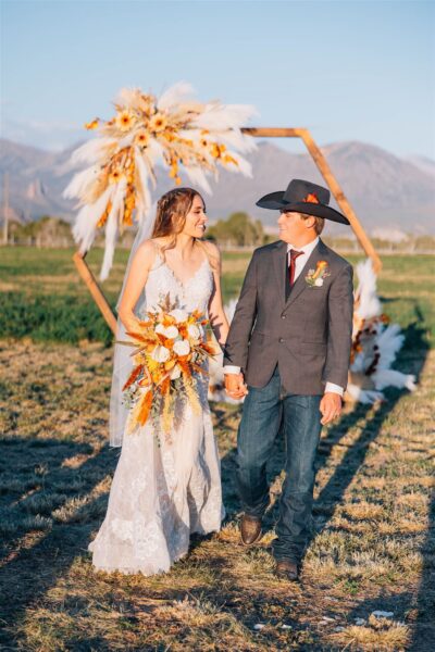Bride and groom holding hands, walking together in a scenic outdoor setting with mountains in the background. The bride wears a lace wedding dress and holds a bouquet with warm autumn colors, while the groom is dressed in a gray suit and cowboy hat. A decorative floral arch is visible behind them, adding to the romantic atmosphere.