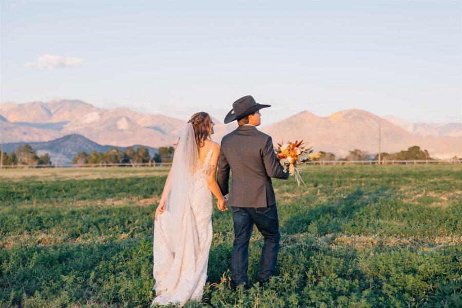 Bride and groom holding hands in a scenic field with mountains in the background, showcasing a romantic outdoor wedding setting. The bride wears a lace wedding gown and veil, while the groom sports a cowboy hat and holds a colorful bouquet.