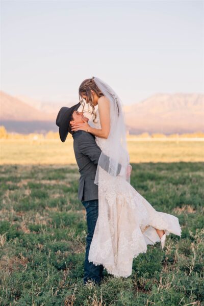 Bride in intricate lace wedding gown being lifted by groom in cowboy hat, set against a scenic mountain backdrop in a grassy field, capturing a romantic moment.