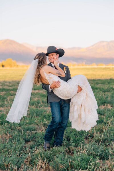 A joyful bride and groom share a romantic moment in a picturesque outdoor setting, with the groom in a cowboy hat and formal attire carrying the bride in a lace wedding dress. The couple is surrounded by lush greenery and mountain scenery in the background, capturing the essence of a rustic wedding celebration.