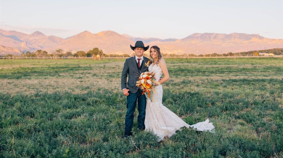 Couple in wedding attire standing in a green field with mountains in the background, the bride wearing a lace dress and holding a colorful bouquet, while the groom is dressed in a suit and cowboy hat. Ideal for showcasing rustic wedding themes and outdoor wedding photography.