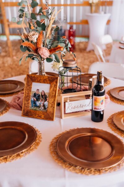 Elegant wedding reception table setup featuring a floral centerpiece, reserved sign, framed photo, and wine bottle, with copper charger plates and a rustic backdrop.