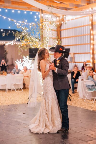 Bride and groom share their first dance at a rustic wedding reception, surrounded by twinkling lights and a warm, romantic atmosphere. The bride wears a stunning lace gown while the groom is dressed in a suit and cowboy hat, creating a charming country vibe. Guests enjoy the celebration in the background.