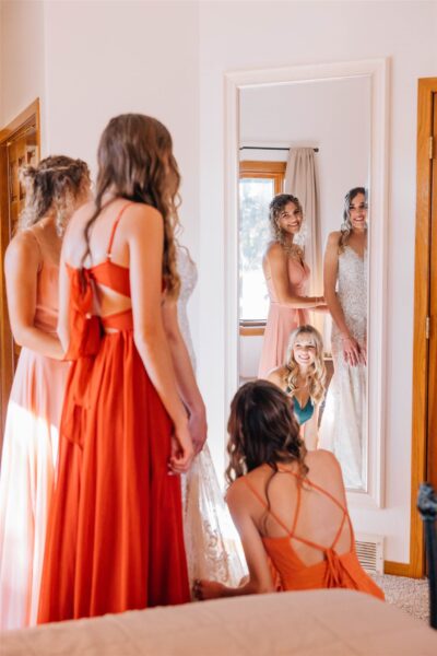 Bridesmaids helping the bride try on her wedding dress in a bright, cozy room with a mirror. The scene captures moments of joy and anticipation, showcasing elegant dresses in various colors.