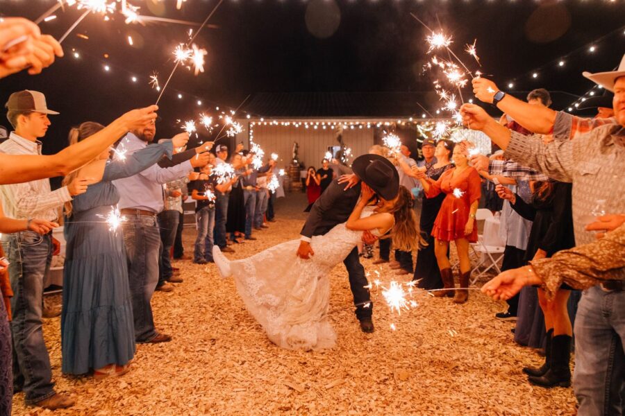 Couple sharing a romantic kiss during their wedding send-off, surrounded by guests holding sparklers under twinkling lights. The scene captures a festive atmosphere with rustic decor and joyful expressions, highlighting a memorable celebration.