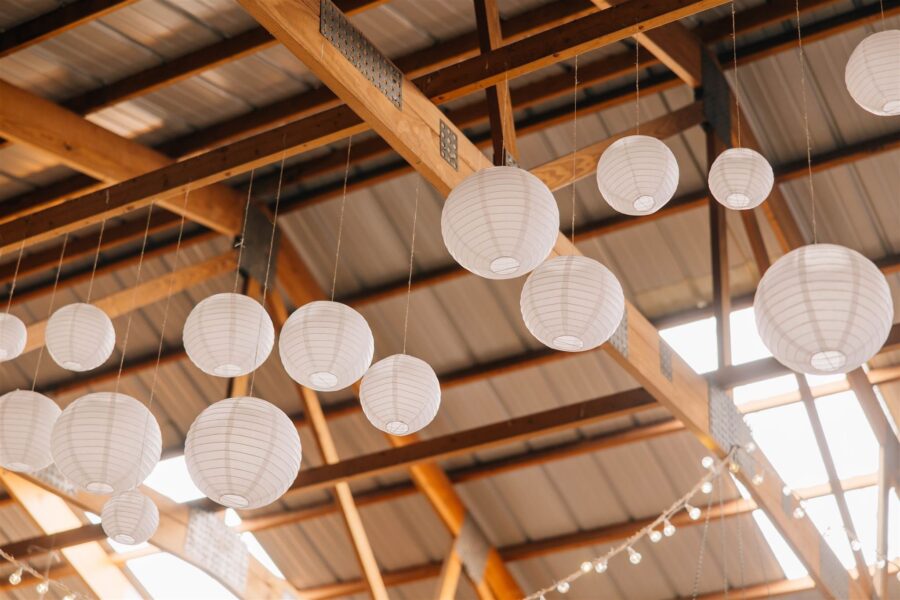 Hanging white paper lanterns create a warm and inviting atmosphere in a rustic venue with wooden beams and a metal roof.