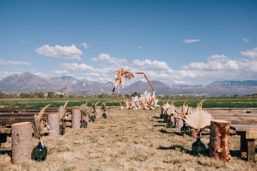 Outdoor wedding ceremony setup featuring wooden benches, decorative vases with dried flowers, and a floral arch against a backdrop of mountains and blue sky. Ideal for rustic and nature-themed weddings.