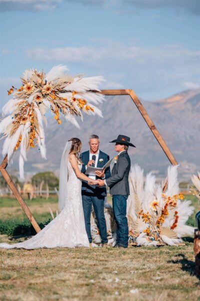 Couple exchanging vows during an outdoor wedding ceremony, framed by a hexagonal wooden arch adorned with pampas grass and autumn flowers, with a scenic mountain backdrop.
