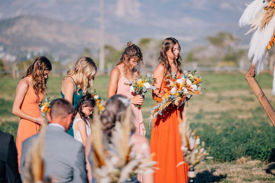 Bridesmaids in colorful dresses, including orange, green, and pink, stand in a field during a wedding ceremony, holding vibrant floral bouquets. A young girl in a white dress is also present, while guests seated in the foreground observe the ceremony against a scenic backdrop.