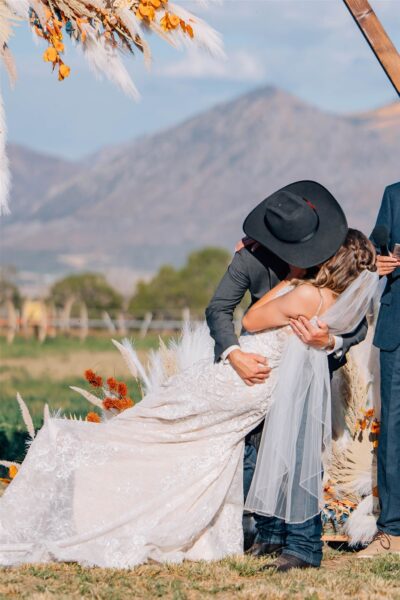 A groom in a black hat lifts his bride, wearing a lace wedding dress and veil, during their outdoor wedding ceremony. The backdrop features decorative floral arrangements and a scenic mountain view, capturing a romantic moment.