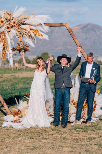 Bride and groom celebrating their wedding ceremony outdoors, surrounded by a floral arch featuring sunflowers and pampas grass, with a celebrant reading from a script nearby. The couple is joyfully raising their arms, showcasing their happiness in a picturesque setting.