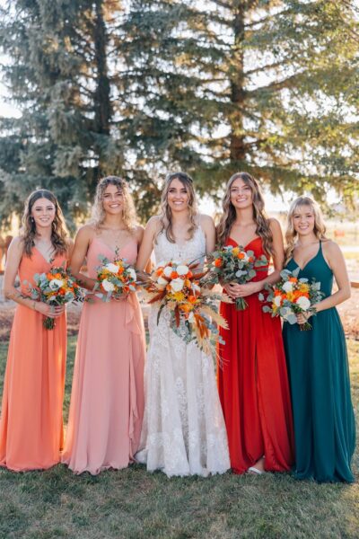 Bridesmaid group photo featuring five women in colorful dresses, standing outdoors against a backdrop of evergreen trees. The bride is in a white lace gown, holding a vibrant bouquet, while each bridesmaid showcases unique floral arrangements with orange and white flowers.